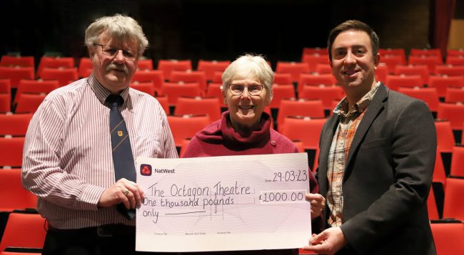 Three people holding a large cheque in front of rows of red seats