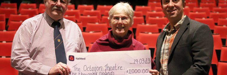 Three people holding a large cheque in front of rows of red seats