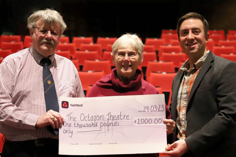 Three people holding a large cheque in front of rows of red seats